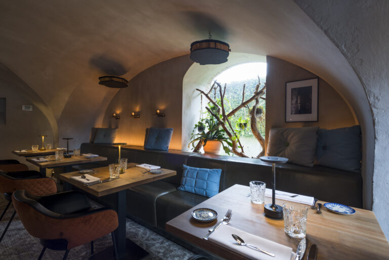 Sfeervol restaurantinterieur met gewelfde muren, houten tafels, leren zitbanken en een raam met weelderig groen.