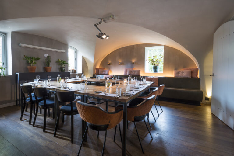 Sfeervolle eetruimte met lange houten tafels, donkere stoelen, oranje fauteuils en een bank met roze kussens onder booggewelven.