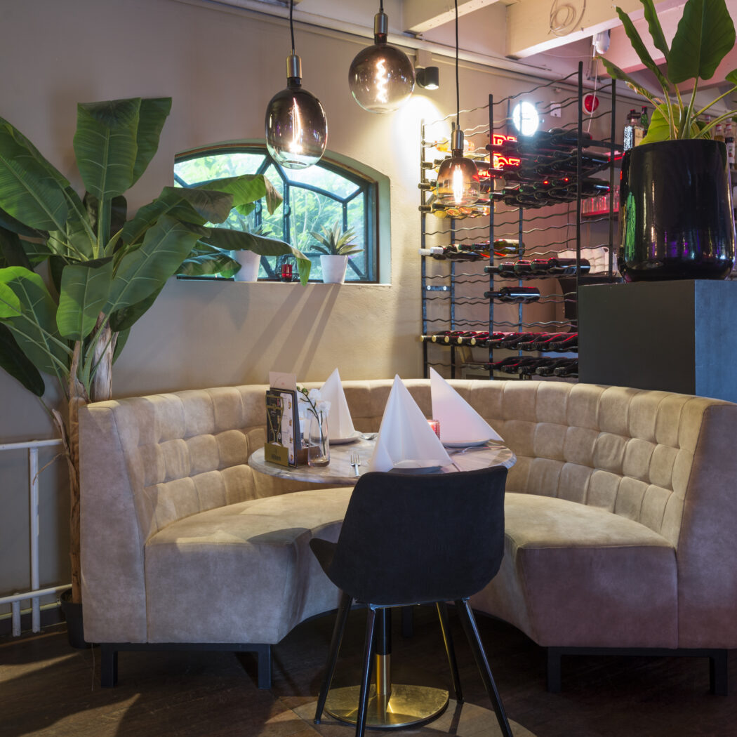 Gezellige zithoek in restaurant met gebogen beige bank, marmeren tafel, planten, wijnrek en moderne lampen.