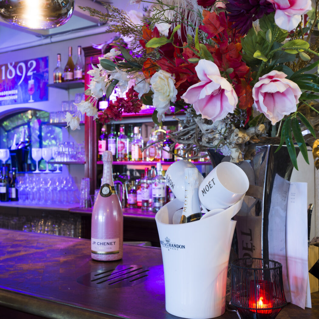 Sfeervolle bar met paarse verlichting, een roze JP Chenet champagne, Moët & Chandon in ijsemmer en groot bloemstuk.