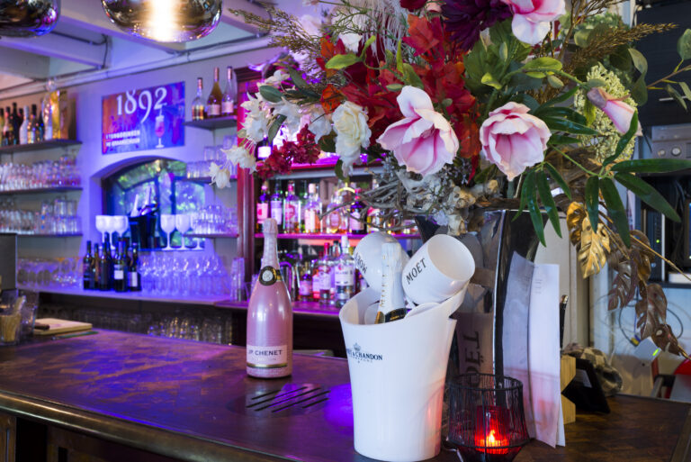 Sfeervolle bar met paarse verlichting, een roze JP Chenet champagne, Moët & Chandon in ijsemmer en groot bloemstuk.