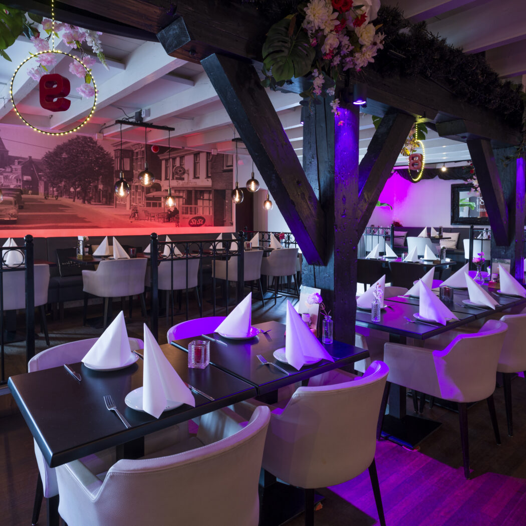 Modern restaurant met gedekte tafels, crèmekleurige stoelen, houten balken en sfeervolle paarse verlichting.