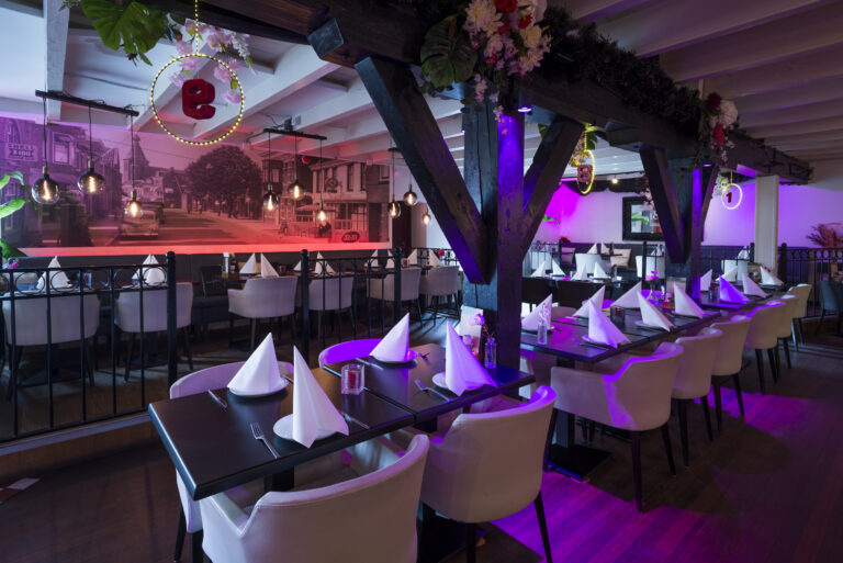 Modern restaurant met gedekte tafels, crèmekleurige stoelen, houten balken en sfeervolle paarse verlichting.