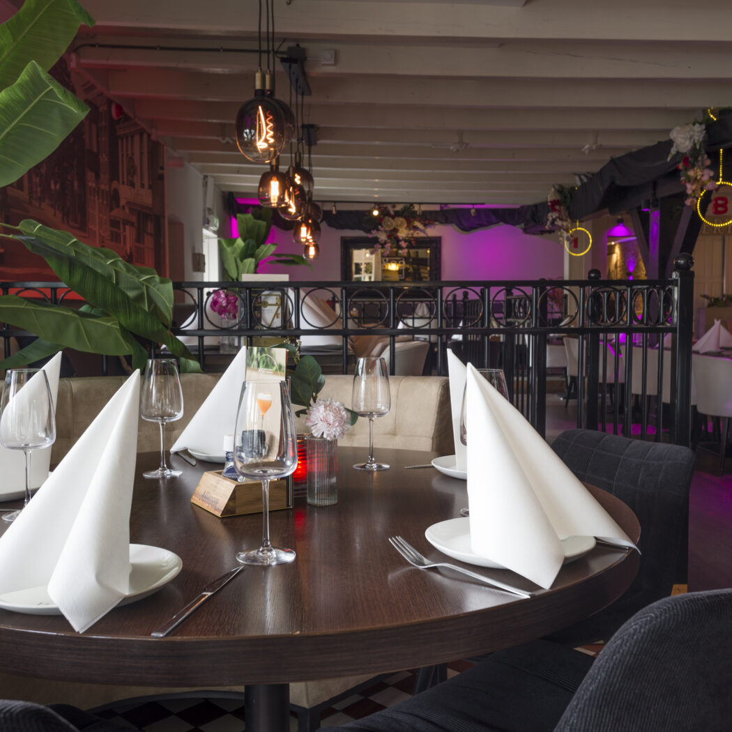 Sfeervol restaurantinterieur met gedekte ronde tafel, paarse verlichting en grote planten.