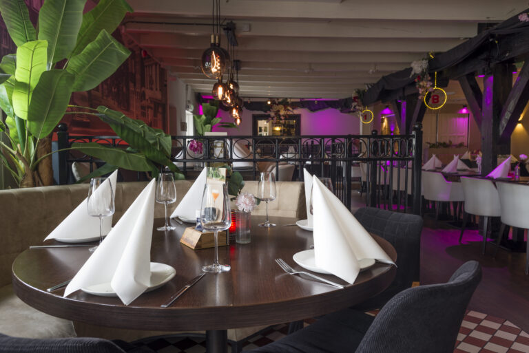 Sfeervol restaurantinterieur met gedekte ronde tafel, paarse verlichting en grote planten.