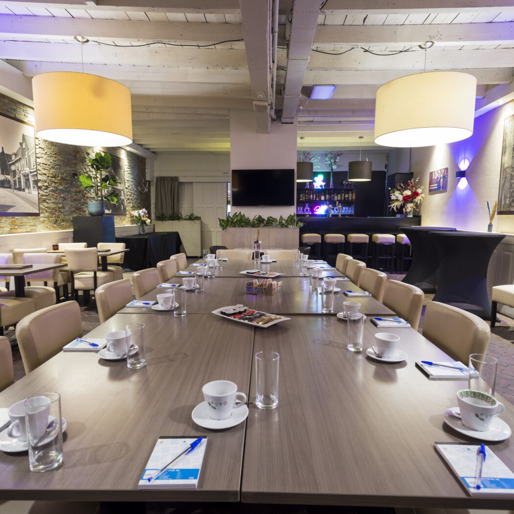 Een lange vergadertafel met beige stoelen, notitieblokken en pennen, in een sfeervolle zaal met bar.