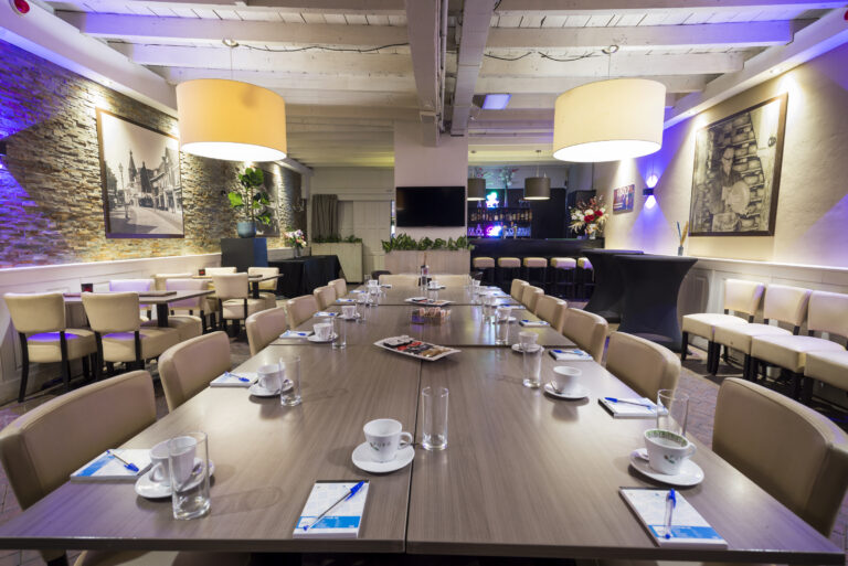 Een lange vergadertafel met beige stoelen, notitieblokken en pennen, in een sfeervolle zaal met bar.