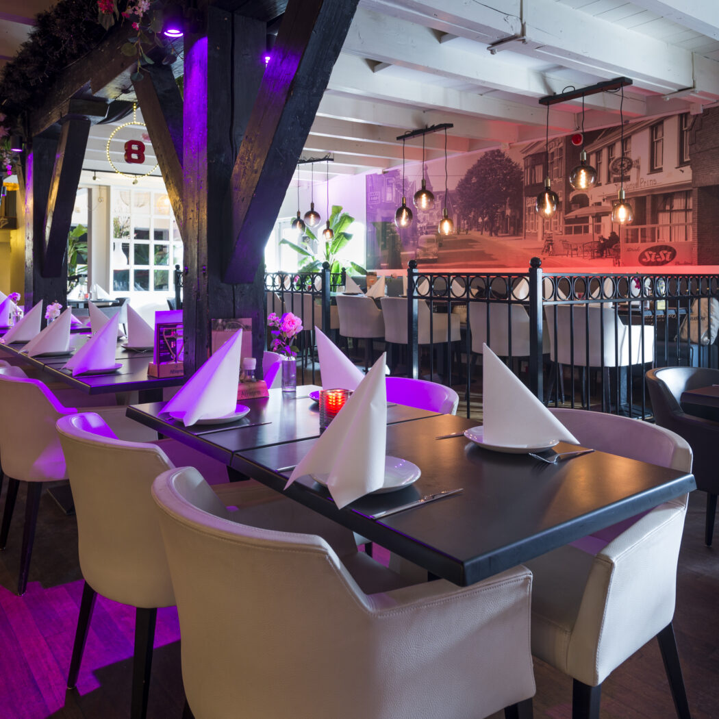 Modern restaurant met lange opgedekte tafels, lichte stoelen, donkere houten balken en paarse sfeerverlichting.