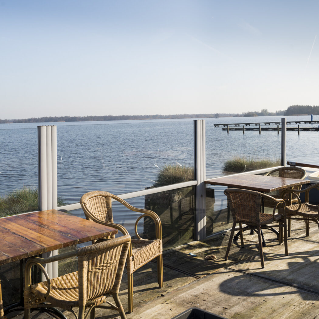 Een zonnig terras aan het water met houten tafels en rieten stoelen, met op de achtergrond een pier en boot.