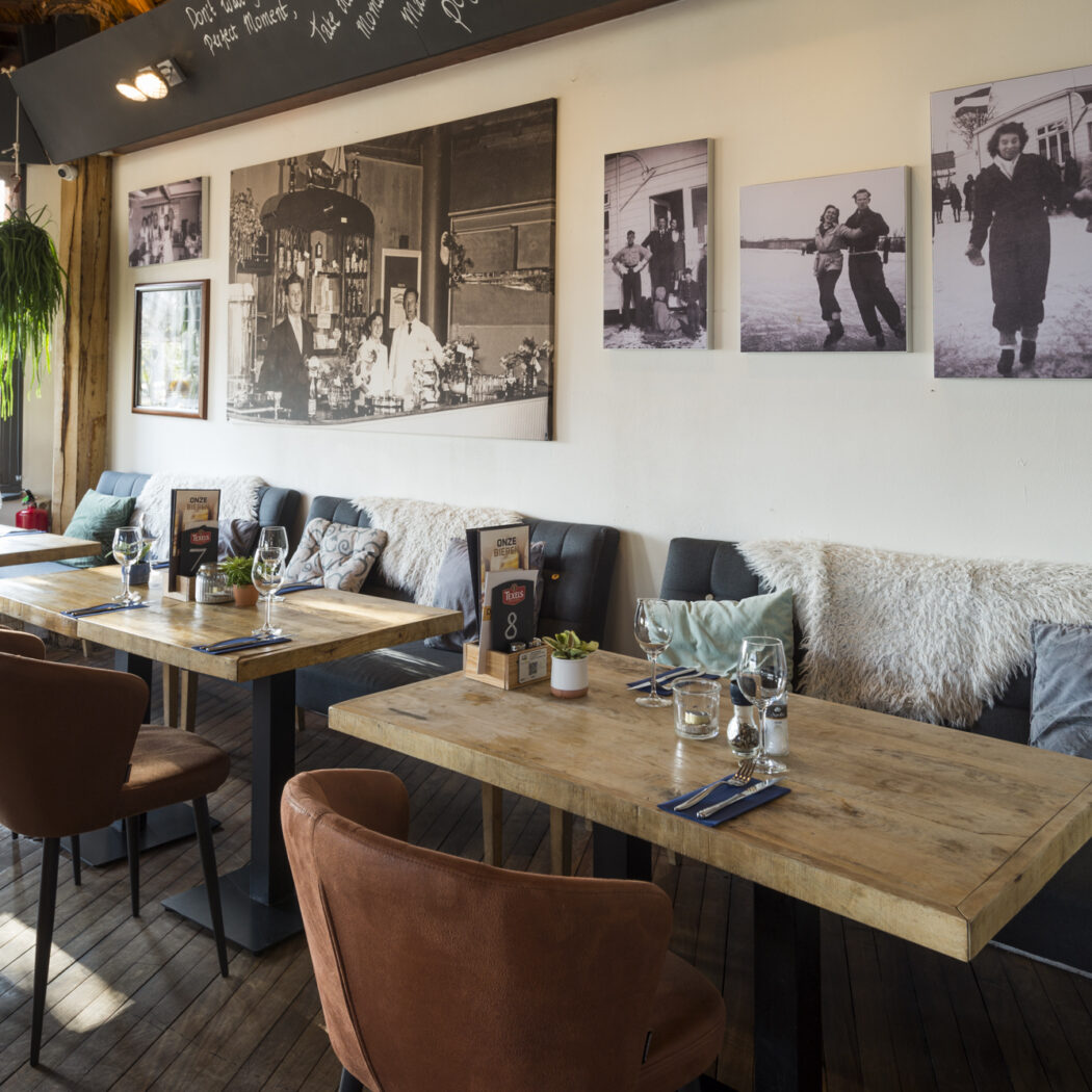 Sfeervolle restaurantinrichting met houten tafels, gestoffeerde banken, bruine stoelen en historische zwart-witfotos aan de wand.