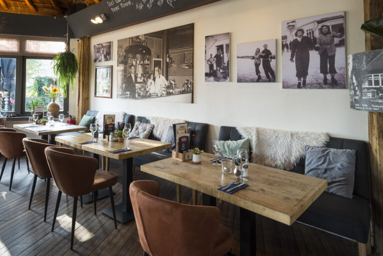 Sfeervolle restaurantinrichting met houten tafels, gestoffeerde banken, bruine stoelen en historische zwart-witfotos aan de wand.