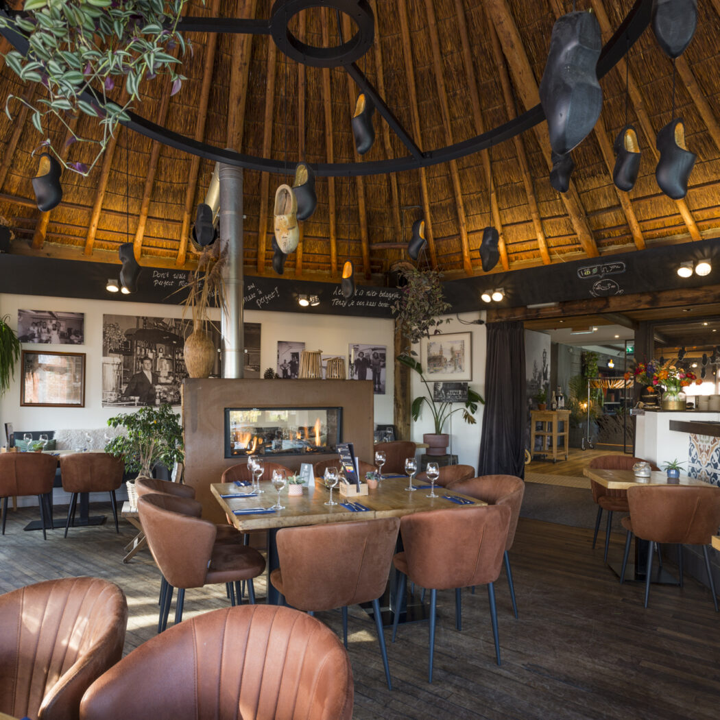 Gezellig restaurant met rieten dak, hangende klompen, open haard en bruine fauteuils aan gedekte tafels.