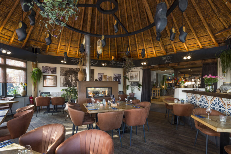 Gezellig restaurant met rieten dak, hangende klompen, open haard en bruine fauteuils aan gedekte tafels.