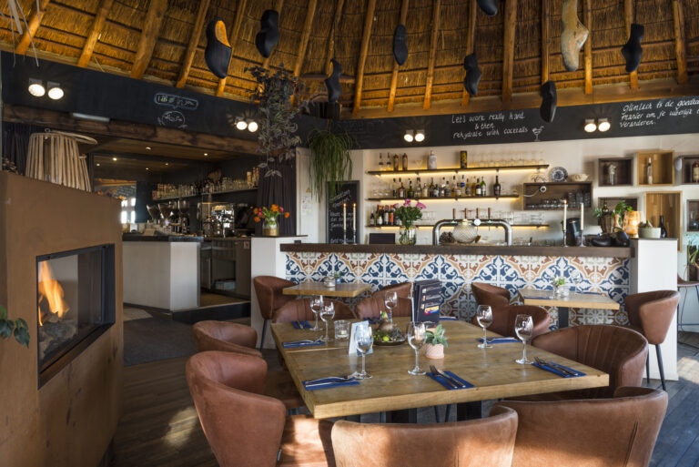 Sfeervol restaurant met rieten dak, een bar met kleurrijke tegels, bruine leren stoelen en een brandende haard.