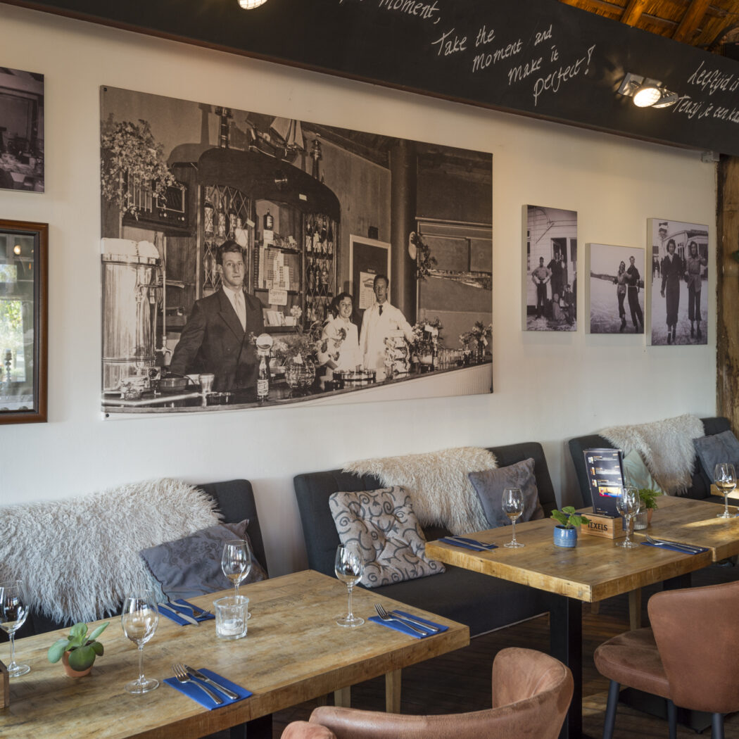 Sfeervol restaurant met gedekte houten tafels, banken, bruine stoelen, groene planten en historische zwart-wit fotos.