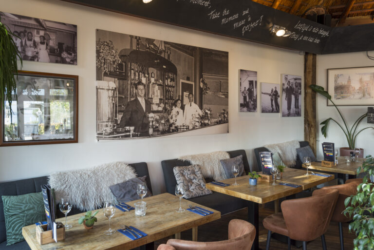 Sfeervol restaurant met gedekte houten tafels, banken, bruine stoelen, groene planten en historische zwart-wit fotos.