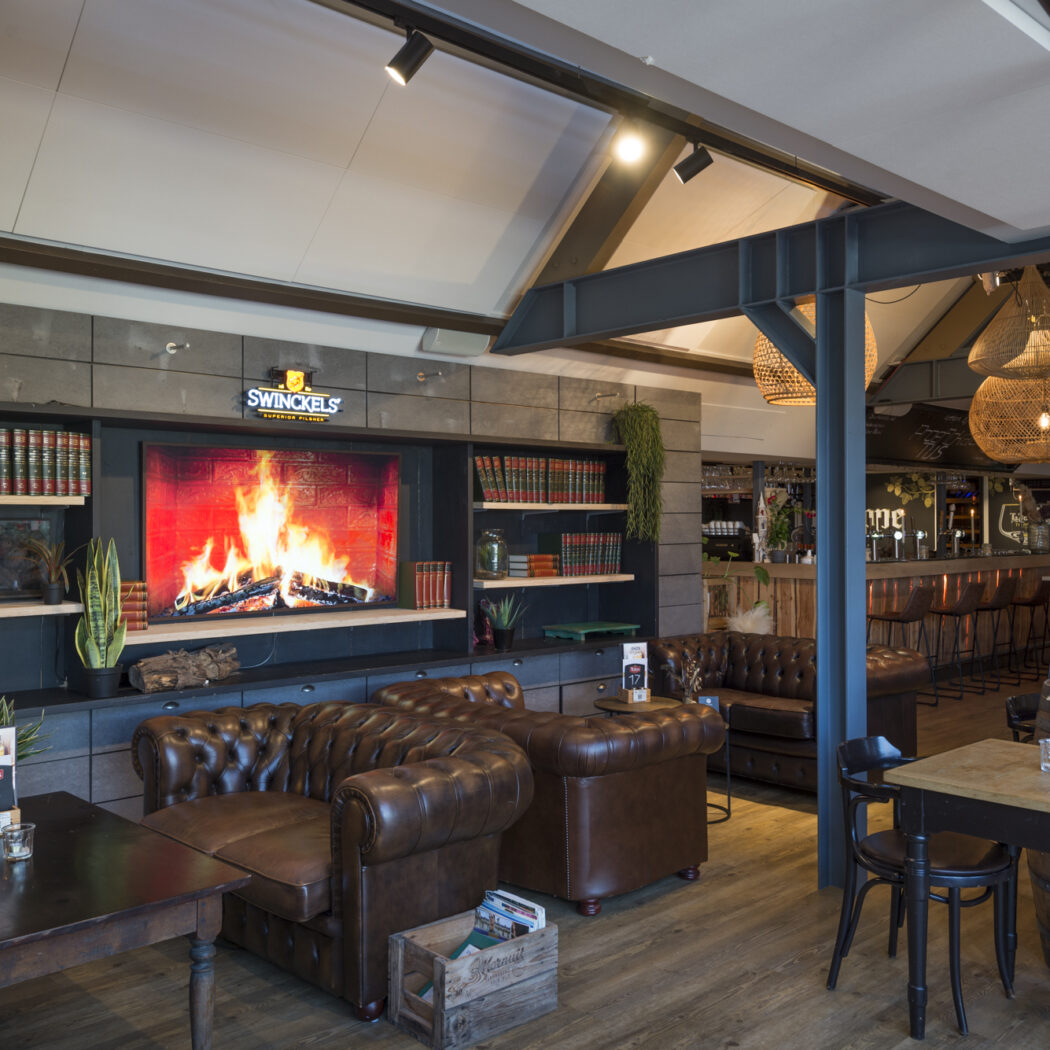 Sfeervol café interieur met een digitale haard, SWINCKELS logo, boekenplanken en bruine Chesterfield banken.
