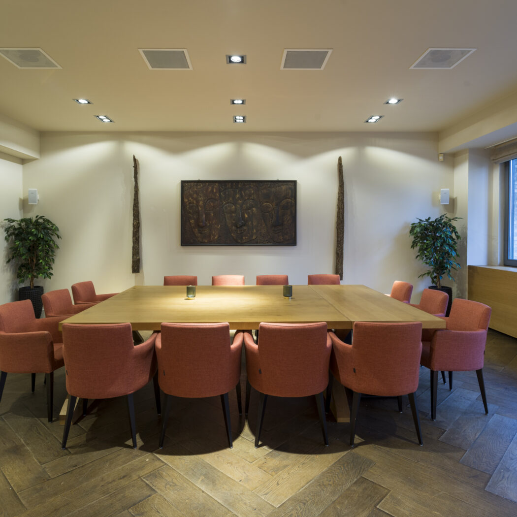 Lichte en warme vergaderzaal met een grote houten tafel, zalmkleurige fauteuils, visgraatvloer, en een donkere wandsculptuur.