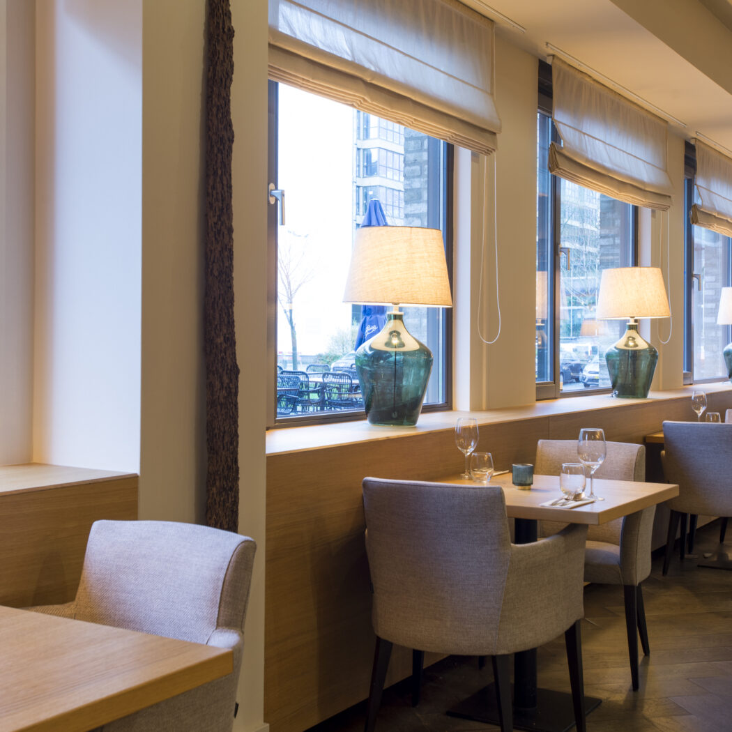 Warme, moderne restaurant eetruimte met houten tafels, grijze stoelen en ramen met blauwgroene lampen.