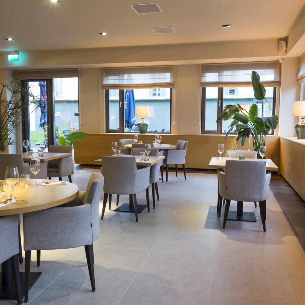 Sfeervolle restaurantzaal met gedekte houten tafels, grijze stoelen en groene lampen bij de ramen.