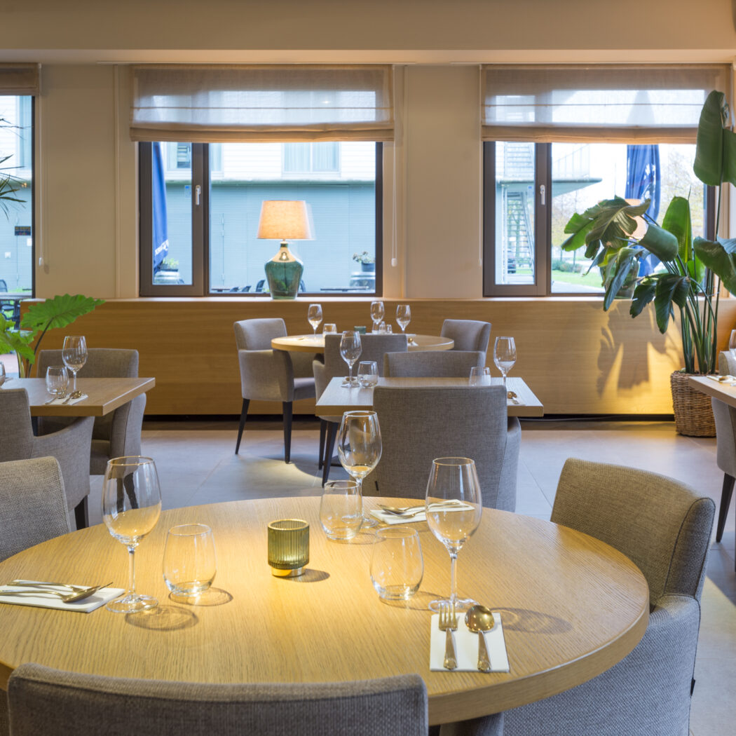 Gedekte houten tafels en grijze stoelen in een sfeervol restaurant met grote ramen, planten en decoratieve lampen.