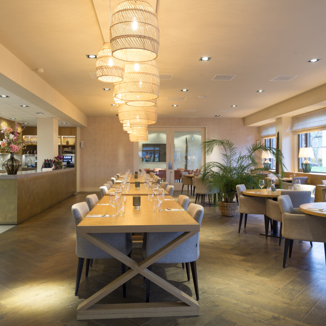 Modern restaurant interieur met houten bar, lange eettafel, ronde tafels en opvallende rieten hanglampen.