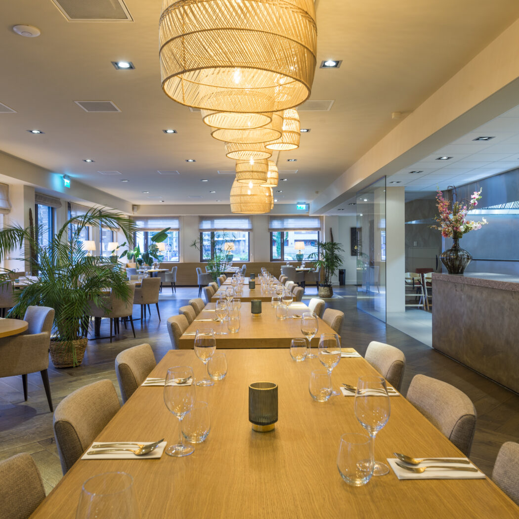 Sfeervol modern restaurantinterieur met lange houten tafels, rotan hanglampen en groene planten, gedekt met bestek en glazen.