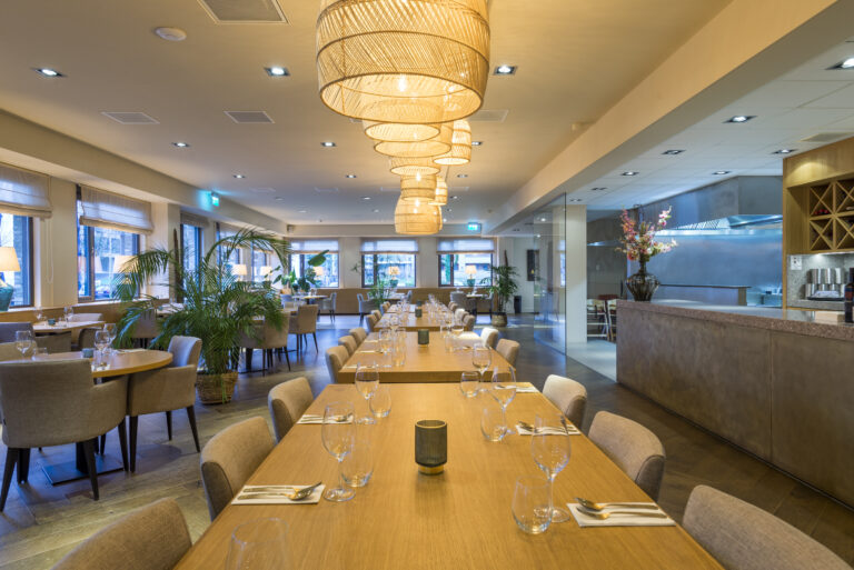 Sfeervol modern restaurantinterieur met lange houten tafels, rotan hanglampen en groene planten, gedekt met bestek en glazen.