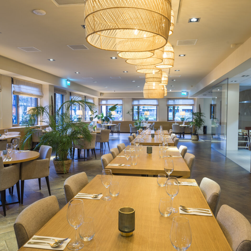 Moderne restaurant eetzaal met lange gedekte houten tafels, rieten hanglampen, planten en grote ramen.