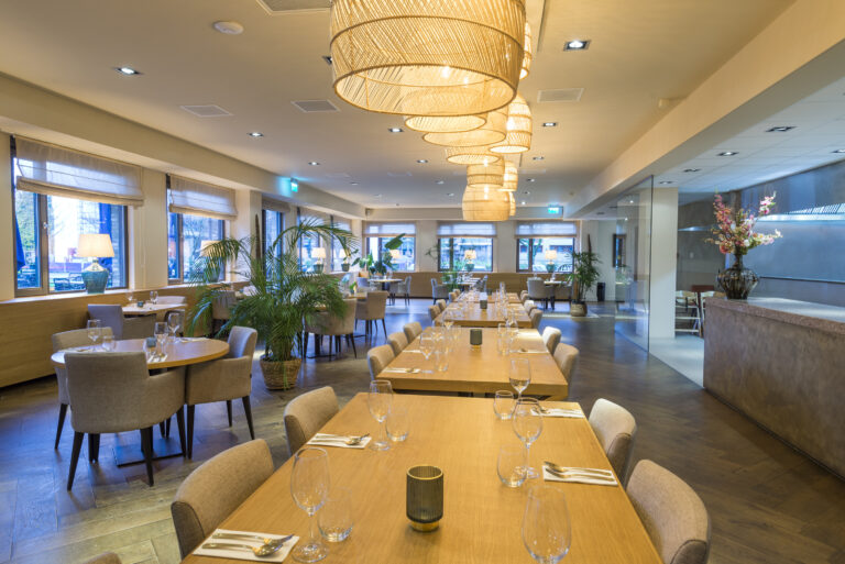 Moderne restaurant eetzaal met lange gedekte houten tafels, rieten hanglampen, planten en grote ramen.