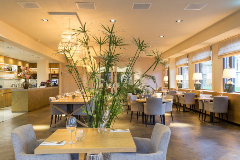 Stijlvol restaurant interieur met houten tafels, grijze stoelen, rieten lampen en weelderige groene planten.
