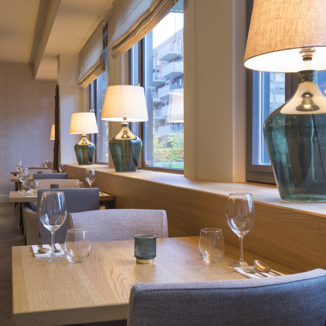 Modern restaurantinterieur met houten tafels, grijze stoelen, gedekte couverts en grote groene glazen lampen bij het raam.