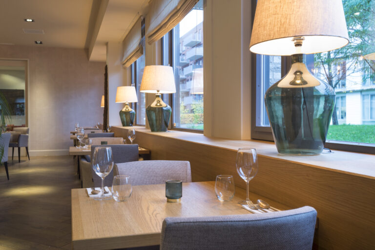 Modern restaurantinterieur met houten tafels, grijze stoelen, gedekte couverts en grote groene glazen lampen bij het raam.