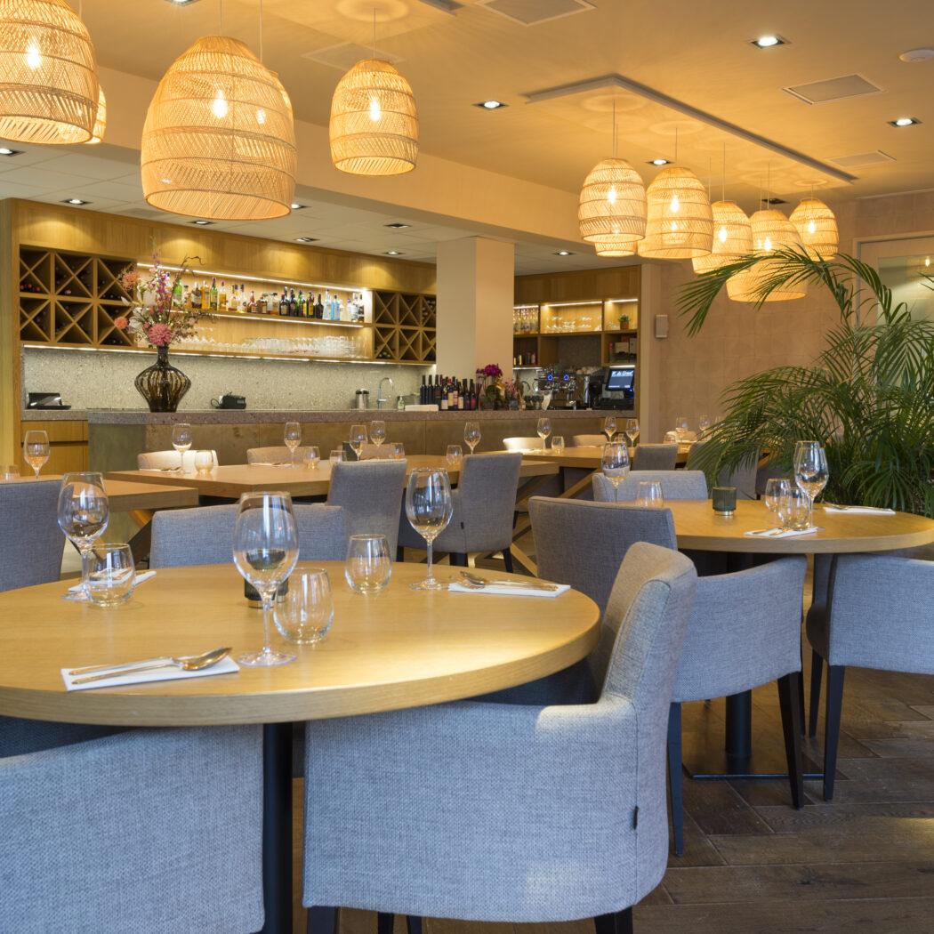 Sfeervol restaurant met ronde houten tafels, grijze stoelen, rieten lampen, een bar en een grote groene plant.