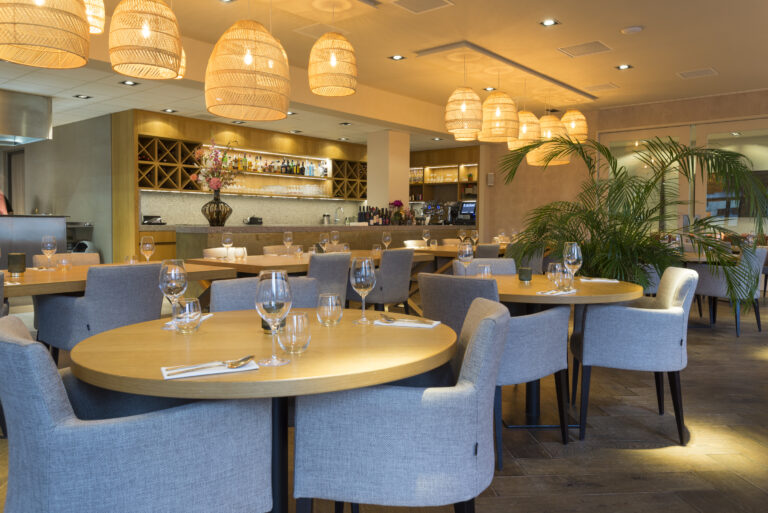 Sfeervol restaurant met ronde houten tafels, grijze stoelen, rieten lampen, een bar en een grote groene plant.