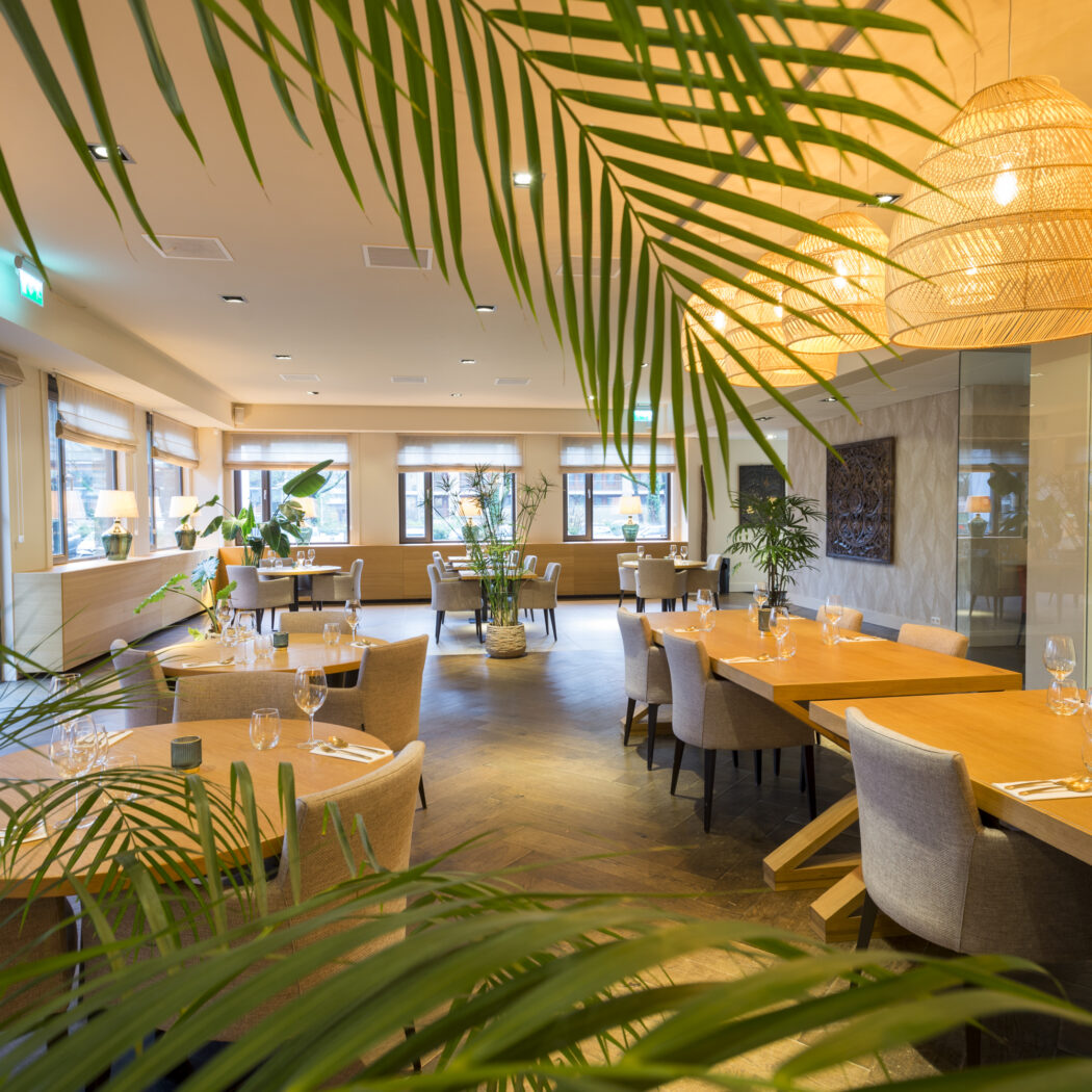 Ruim, licht restaurant met gedekte houten tafels, groene planten en rieten lampen, omkaderd door palmbladeren.