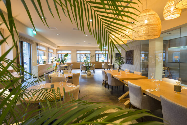 Ruim, licht restaurant met gedekte houten tafels, groene planten en rieten lampen, omkaderd door palmbladeren.