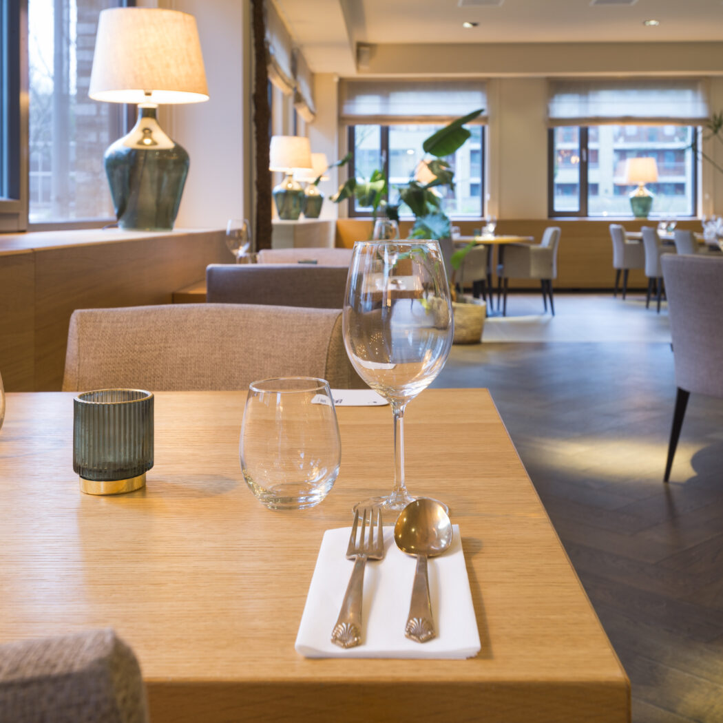 Gedekte houten tafel met wijnglas en bestek in een stijlvol restaurant met sfeervolle verlichting.