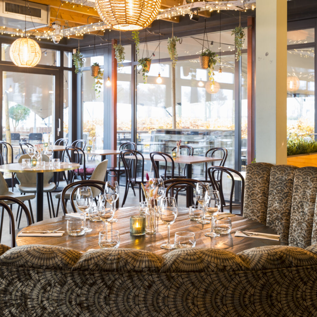 Sfeervol restaurant met een ronde houten eettafel, patroonbank, rieten lampen en grote ramen met zicht naar buiten.
