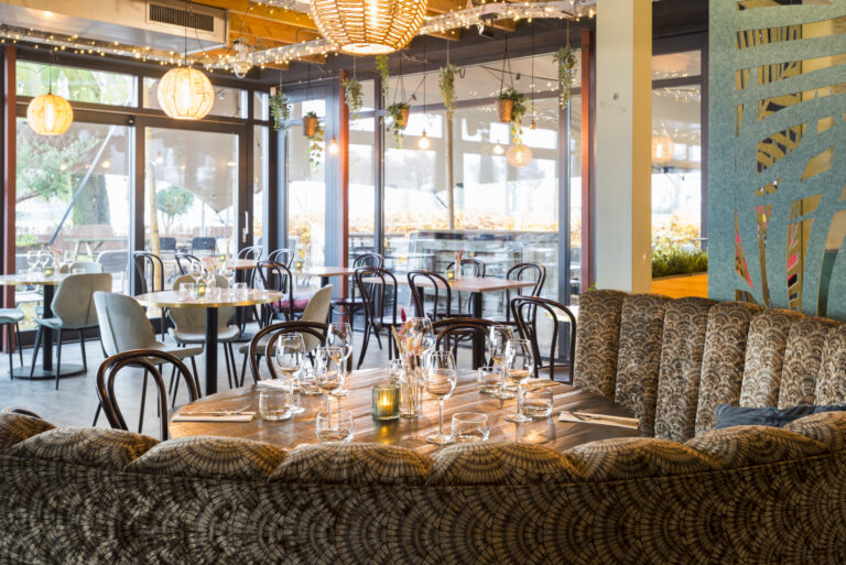 Sfeervol restaurant met een ronde houten eettafel, patroonbank, rieten lampen en grote ramen met zicht naar buiten.
