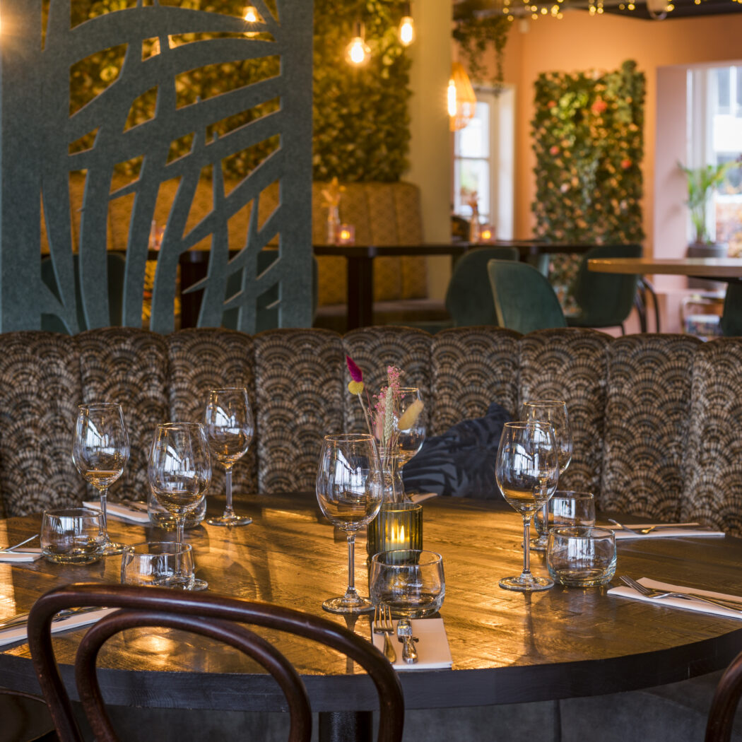 Sfeervol restaurantinterieur met gedekte houten tafel, gestoffeerde patroonbank en groene plantenmuren.
