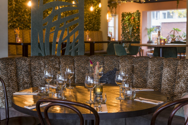 Sfeervol restaurantinterieur met gedekte houten tafel, gestoffeerde patroonbank en groene plantenmuren.