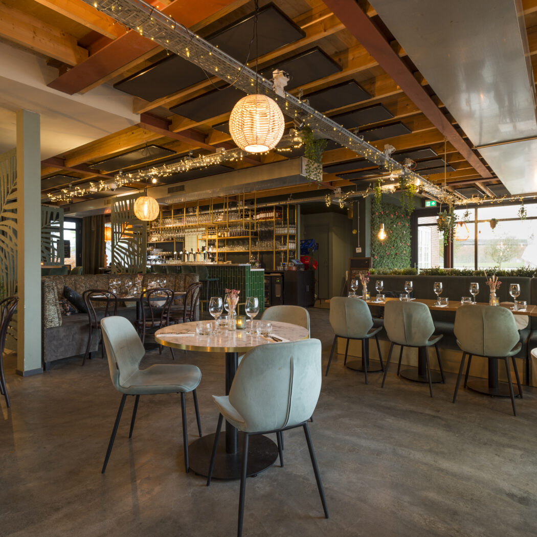 Stijlvol restaurantinterieur met houten balken, groene banken, bladmotief panelen en een verlichte bar.
