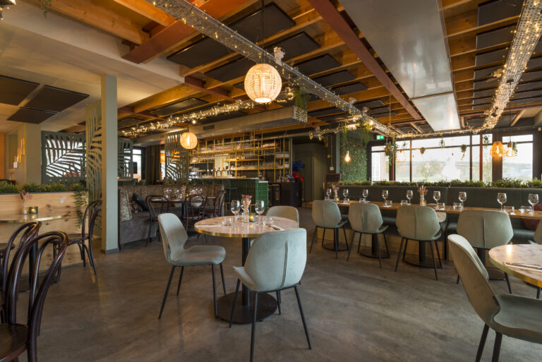 Stijlvol restaurantinterieur met houten balken, groene banken, bladmotief panelen en een verlichte bar.
