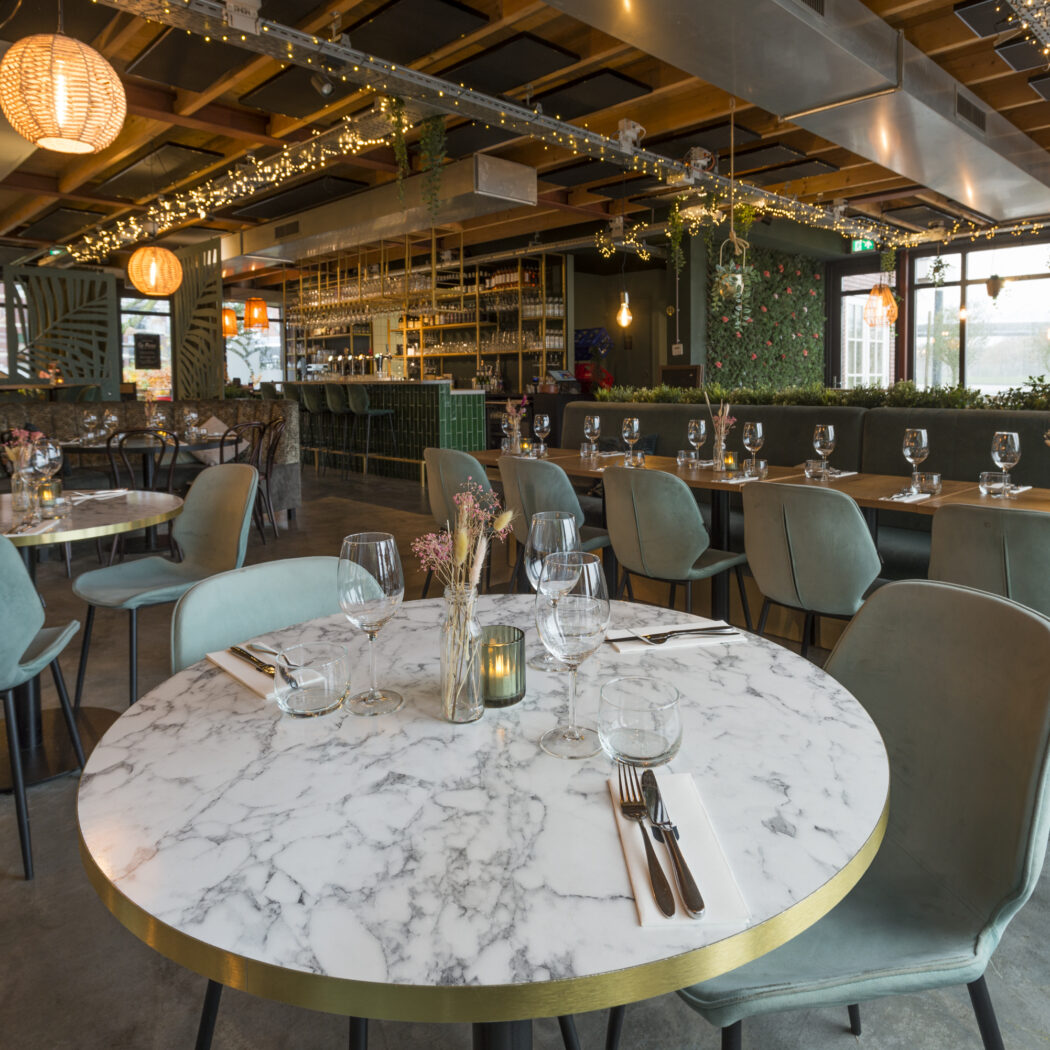Sfeervol modern restaurantinterieur met ronde marmeren tafels, zachtgroene stoelen, een bar en warme sfeerverlichting.