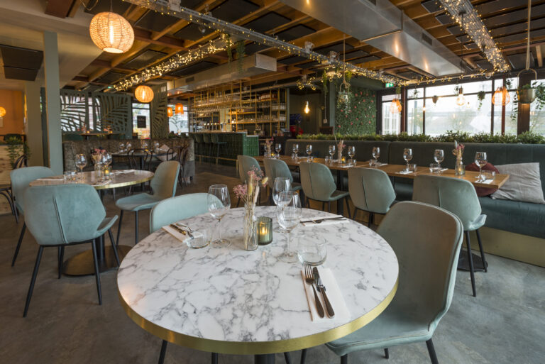 Sfeervol modern restaurantinterieur met ronde marmeren tafels, zachtgroene stoelen, een bar en warme sfeerverlichting.