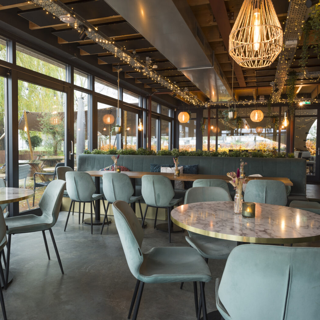 Sfeervol restaurant met lichtgroene fluwelen stoelen, marmeren tafels en hanglampen bij grote ramen.