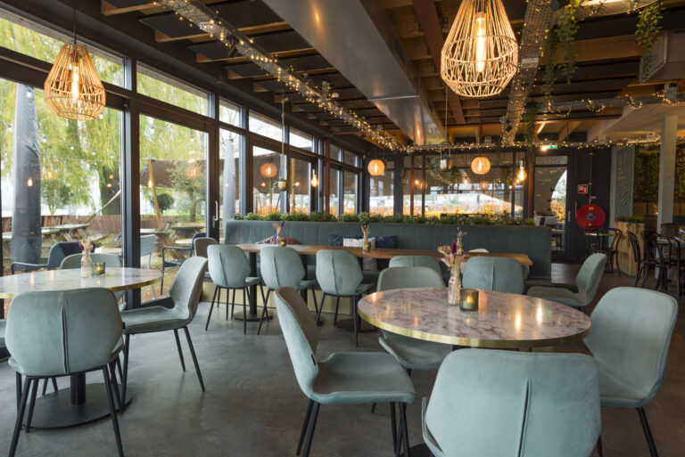Sfeervol restaurant met lichtgroene fluwelen stoelen, marmeren tafels en hanglampen bij grote ramen.