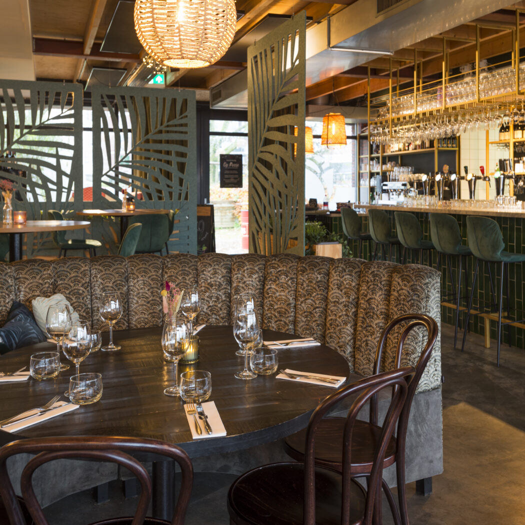 Sfeervol restaurantinterieur met gedekte ronde tafel, banketbank, groene schermen en een stijlvolle bar.