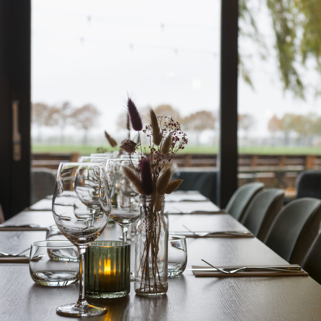Lange gedekte restauranttafel met wijnglazen, bestek, droogbloemen en kaarslicht, uitzicht op bomenrij.